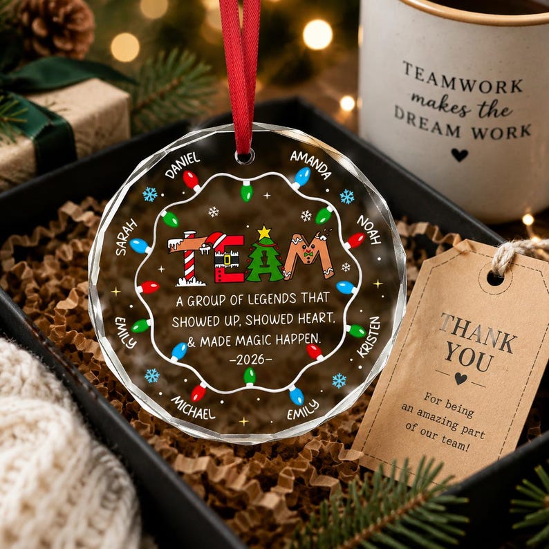 May include: A round, clear glass Christmas ornament with a red ribbon hanger. The design features a string of colorful holiday lights, the word TEAM, and the text: A group of legends that showed up, showed heart, & made magic happen. -2026-