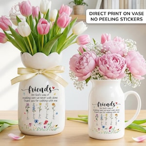 May include: Two white ceramic vases, one with a scalloped rim and a bow, the other with a handle, each holding flowers. The vases are decorated with the word "friends" and a floral design. One vase holds pink and white tulips, the other holds pink peonies.