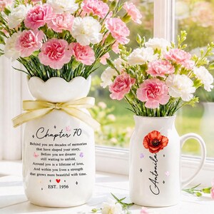 May include: A white ceramic vase with a scalloped rim and a yellow ribbon, featuring the text Chapter 70 and a poem, alongside a white ceramic mug with a red poppy graphic and the name Antonia, both filled with pink and white carnations.