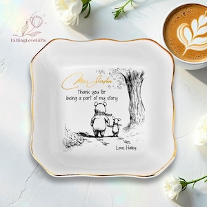 May include: A white ceramic trinket dish with a gold trim. The dish features a black and white illustration of a bear and a pig walking together, with the text "Miss Jorden, thank you for being a part of my story, Love, Hailey."
