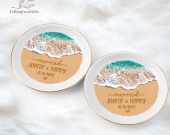 May include: Two round white ceramic trinket dishes with gold-toned rims. Each dish features a printed design of a sandy beach with turquoise ocean waves and white sea foam. The text reads married JENIFER & HARRY 10.10.2024 with a small heart.