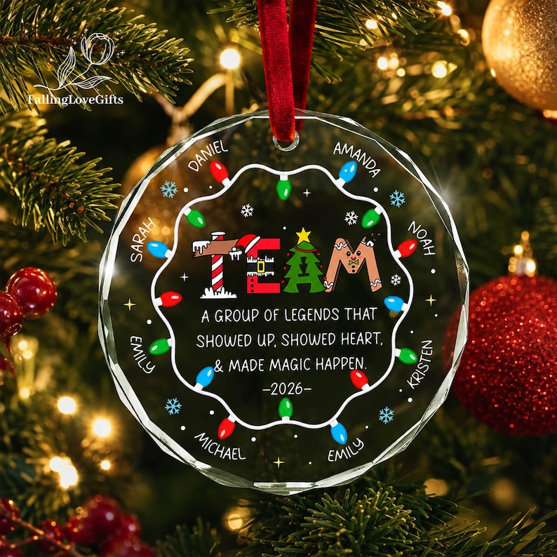 May include: A round, clear glass Christmas ornament with a red velvet ribbon. The design features the word TEAM in festive letters, surrounded by a string of colorful lights and snowflakes. It includes the names Daniel, Amanda, Noah, Kristen, Emily, Michael, Sarah, and Emily, with the text: A GROUP OF LEGENDS THAT SHOWED UP, SHOWED HEART, & MADE MAGIC HAPPEN. -2026-.