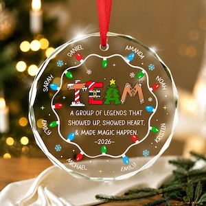 May include: A round, clear glass Christmas ornament featuring a red ribbon hanger. The design includes the word TEAM in festive letters, a string of colorful holiday lights, and the text: A GROUP OF LEGENDS THAT SHOWED UP, SHOWED HEART, & MADE MAGIC HAPPEN. -2026- with names Daniel, Amanda, Noah, Kristen, Emily, Michael, and Sarah.