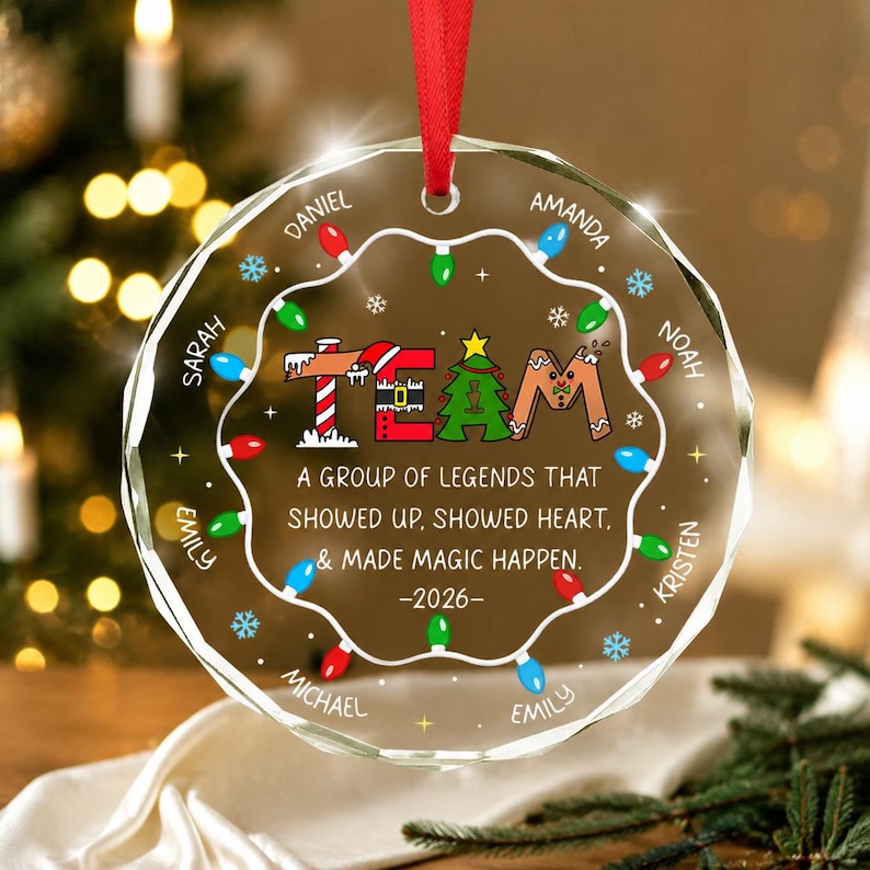 May include: A round, clear glass Christmas ornament featuring a red ribbon hanger. The design includes the word TEAM in festive letters, a string of colorful holiday lights, and the text: A GROUP OF LEGENDS THAT SHOWED UP, SHOWED HEART, & MADE MAGIC HAPPEN. -2026- with names Daniel, Amanda, Noah, Kristen, Emily, Michael, and Sarah.