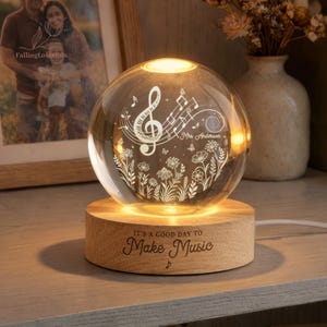Pu&ograve; includere: Una luce notturna a sfera di cristallo luminosa con una base in legno. La sfera presenta un design a note musicali e il testo "It's a good day to Make Music". La luce &egrave; su un tavolo di legno.