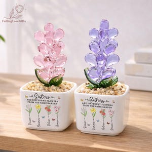 May include: Two square white ceramic planters containing glass flower sculptures. One features pink glass petals, the other purple. Both pots display the text "Sisters are different flowers from the same garden" with floral illustrations and names.