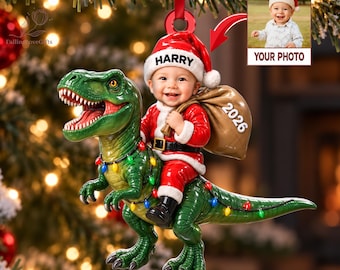 May include: A 3D acrylic Christmas ornament featuring a green Tyrannosaurus Rex decorated with colorful string lights, carrying a baby dressed as Santa Claus. The baby wears a hat with the name HARRY and holds a sack labeled 2026.