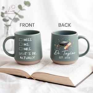 May include: A teal ceramic mug with a tan base, featuring text on both sides. The front says "MISS, MS., MRS., IT'S DR. ACTUALLY." The back shows a graduation cap, diploma, and "Dr. Taylor EST. 2026."