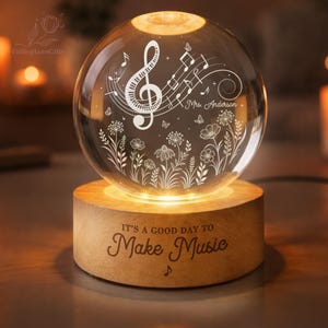 Pu&ograve; includere: Una sfera di vetro illuminata con note musicali e motivi floreali, appoggiata su una base di legno. La base recita "IT'S A GOOD DAY TO Make Music". La sfera si illumina dall'interno, creando un'atmosfera calda e invitante.