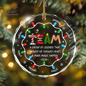 May include: A circular clear glass Christmas ornament featuring a string of colorful holiday lights around the perimeter. The center displays the word TEAM with festive graphics, the text A GROUP OF LEGENDS THAT SHOWED UP, SHOWED HEART, & MADE MAGIC HAPPEN. -2026-, and several names.