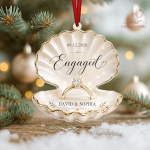 May include: A cream-colored, shell-shaped ceramic ornament with gold trim, hanging from a red ribbon. It features the text 09.12.2026, Engaged, and DAVID & SOPHIA, with a central gold ring illustration. The design includes small decorative leaf accents.