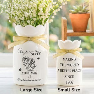 May include: Two white ceramic vases with scalloped rims and gold ribbon accents. The larger vase has text that reads "Chapter SIXTY" and "STEPHANIE". The smaller vase says "MAKING THE WORLD A BETTER PLACE SINCE 1966". Both vases are filled with flowers.