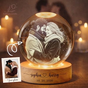 May include: A spherical glass ornament featuring an etched image of a couple embracing, set on a circular wooden base. The base is engraved with the names Sophia and Harry, a heart symbol, and the date 10. 20. 2026. A small photo print is included.