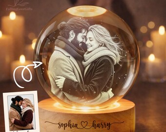 Può includere: Un ornamento sferico in vetro con l'immagine incisa di una coppia che si abbraccia, posto su una base circolare in legno. La base è incisa con i nomi Sophia e Harry, un simbolo a cuore e la data 10. 20. 2026. È inclusa una piccola foto.