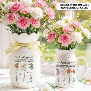 May include: Two white ceramic vases with floral arrangements. One vase has a scalloped top and a yellow ribbon. Both vases have the text "Sisters are different flowers from the same garden" and names. The flowers are pink and white carnations.