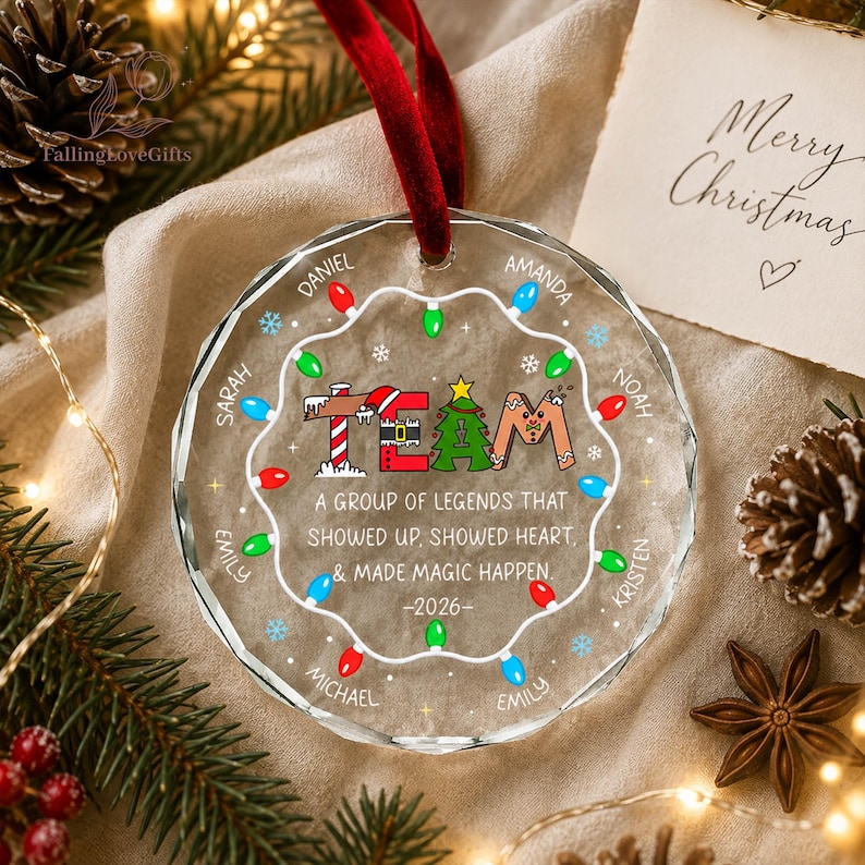 May include: A round, faceted glass Christmas ornament hanging from a red velvet ribbon. The ornament features a string of colorful lights, the word TEAM, and the text: A GROUP OF LEGENDS THAT SHOWED UP, SHOWED HEART, & MADE MAGIC HAPPEN. -2026-