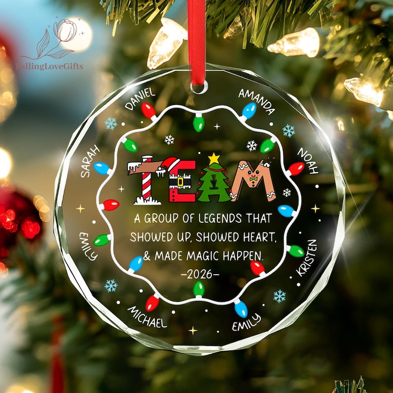 May include: A round, clear glass Christmas ornament with a red ribbon hanger. The design features a string of colorful lights, snowflakes, and the word TEAM with festive graphics. It includes the text A GROUP OF LEGENDS THAT SHOWED UP, SHOWED HEART, & MADE MAGIC HAPPEN. -2026- and several names arranged in a circle.