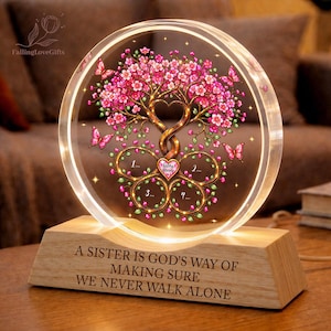 May include: A circular acrylic plaque featuring a printed tree with pink blossoms and butterflies, set on a light wood base. The tree trunk forms a heart shape with four circular frames labelled 1, 2, 3, and 4. The base is engraved with the text: A SISTER IS GOD'S WAY OF MAKING SURE WE NEVER WALK ALONE.