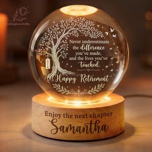 May include: A spherical glass ornament on a round wooden base. The glass features an etched tree, birds, and the text: Never underestimate the difference you've made, and the lives you've touched. Happy Retirement. The base reads: Enjoy the next chapter Samantha.