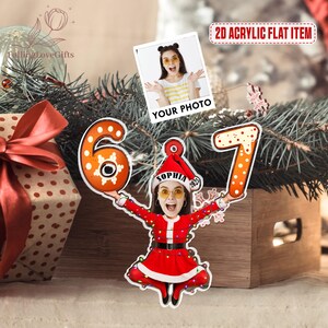 May include: A personalized Christmas ornament featuring a photo and the numbers 6 and 7. The ornament shows a person in a Santa Claus outfit holding the numbers. The text "YOUR PHOTO" is above the photo. The text "2D ACRYLIC FLAT ITEM" is in the upper right corner.