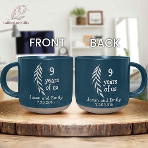 May include: A pair of teal ceramic mugs with white text and a leaf design. Each mug reads "9 years of us," with the names "Jason and Emily" and the date "7.25.2016." The mugs have a light gray base and are displayed on a wooden tray.