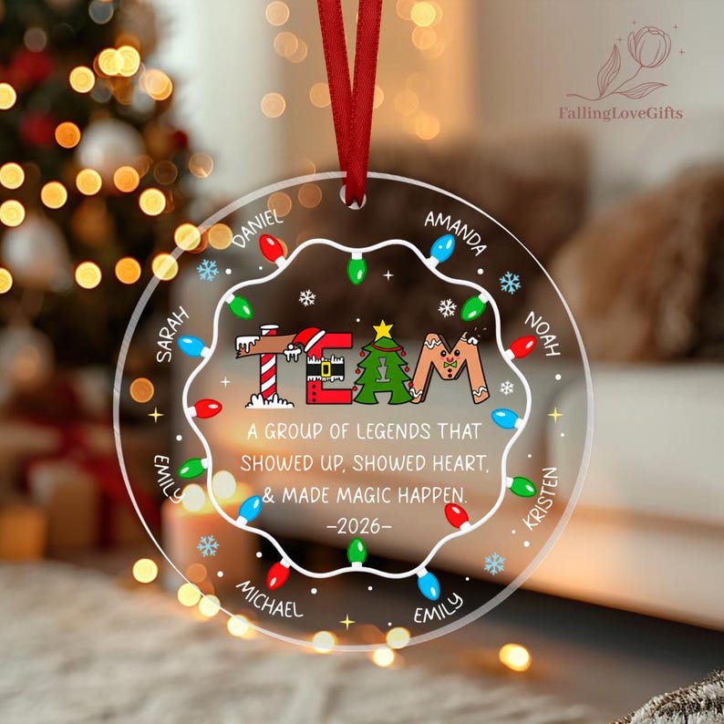 May include: A clear circular acrylic Christmas ornament hanging from a red ribbon. It features a string of colorful holiday lights, the word TEAM, and the text: A GROUP OF LEGENDS THAT SHOWED UP, SHOWED HEART, & MADE MAGIC HAPPEN. -2026-. Names are printed around the edge.