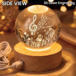 Pu&ograve; includere: Una sfera di cristallo trasparente con incisione laser 3D di note musicali, fiori e la scritta "Mrs. Anderson". La sfera poggia su una base di legno che emette una calda luce. I testi "SIDE VIEW" e "3D Laser Engraving" sono visibili.