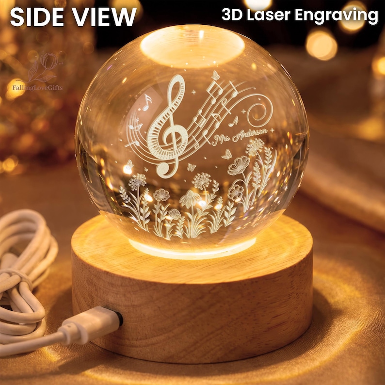 Pu&ograve; includere: Una sfera di cristallo trasparente con incisione laser 3D di note musicali, fiori e la scritta "Mrs. Anderson". La sfera poggia su una base di legno che emette una calda luce. I testi "SIDE VIEW" e "3D Laser Engraving" sono visibili.