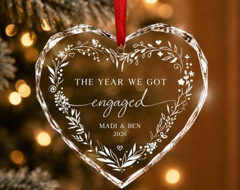 May include: A heart-shaped clear glass ornament with faceted edges, featuring a white etched floral wreath design. The text reads THE YEAR WE GOT engaged, MADI & BEN, and 2026. A red ribbon is attached to the top for hanging on a tree.