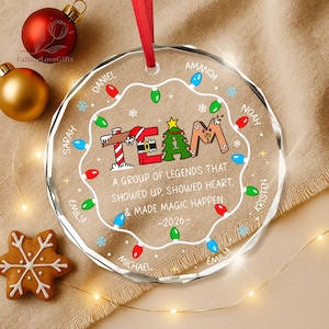 May include: A round, clear acrylic Christmas ornament with a red ribbon hanger. The design features a string of colorful holiday lights, a gingerbread-style 'TEAM' graphic, and the text 'A GROUP OF LEGENDS THAT SHOWED UP, SHOWED HEART, & MADE MAGIC HAPPEN -2026-' with several names printed around the border.
