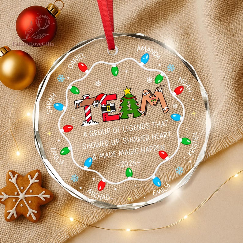 May include: A round, clear acrylic Christmas ornament with a red ribbon hanger. The design features a string of colorful holiday lights, a gingerbread-style 'TEAM' graphic, and the text 'A GROUP OF LEGENDS THAT SHOWED UP, SHOWED HEART, & MADE MAGIC HAPPEN -2026-' with several names printed around the border.