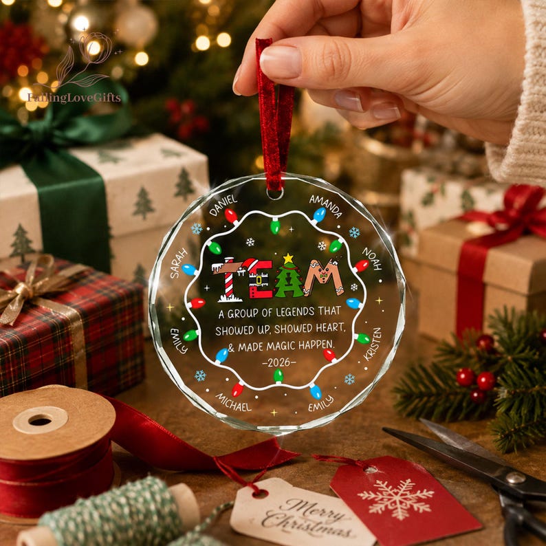May include: A round, clear glass Christmas ornament featuring a red ribbon hanger. The design includes a string of colorful holiday lights, a Christmas tree, and the text: A GROUP OF LEGENDS THAT SHOWED UP, SHOWED HEART, & MADE MAGIC HAPPEN. -2026-
