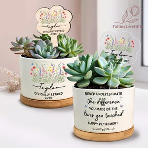 May include: Two white ceramic succulent planters with wooden bases. One planter has a wooden tag that reads "Taylor Officially Retired -2026-". The other planter reads "Never underestimate the difference you made or the lives you touched. Happy Retirement."