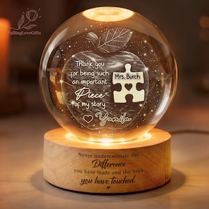 May include: A lit crystal sphere with an apple design and the words "Thank you for being such an important piece of my story." The sphere sits on a wooden base with the text "Never underestimate the difference you have made and the lives you have touched."