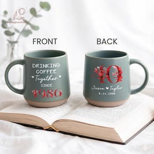 May include: Two teal ceramic mugs with a tan base are displayed on an open book. The front mug reads "DRINKING COFFEE Together SINCE 1986". The back mug has the number "40" with floral accents, the names "Jason & Taylor" and the date "6.11.1986".