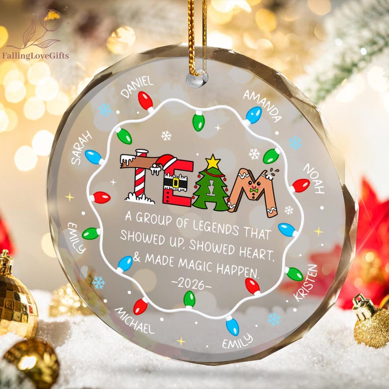May include: A round, clear glass Christmas ornament featuring a string of colorful holiday lights around the edge. The center displays the word TEAM with festive graphics, the text A GROUP OF LEGENDS THAT SHOWED UP, SHOWED HEART, & MADE MAGIC HAPPEN. -2026-, and several names.
