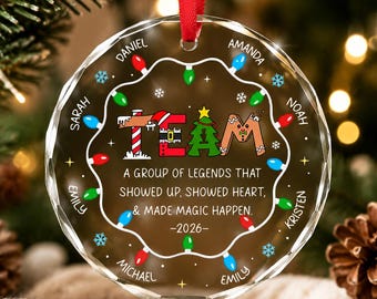 May include: A round, clear glass Christmas ornament with a red ribbon hanger. The design features a string of colorful holiday lights, snowflakes, and the text: TEAM A GROUP OF LEGENDS THAT SHOWED UP, SHOWED HEART, & MADE MAGIC HAPPEN. -2026-