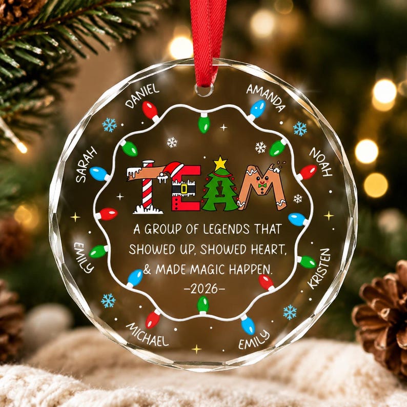 May include: A round, clear glass Christmas ornament with a red ribbon hanger. The design features a string of colorful holiday lights, snowflakes, and the text: TEAM A GROUP OF LEGENDS THAT SHOWED UP, SHOWED HEART, & MADE MAGIC HAPPEN. -2026-
