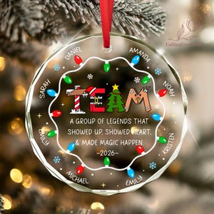 May include: A round, clear glass Christmas ornament with a red ribbon hanger. It features a string of colorful holiday lights, snowflakes, and the text "TEAM" with festive icons. It includes the quote "A GROUP OF LEGENDS THAT SHOWED UP, SHOWED HEART, & MADE MAGIC HAPPEN. -2026-" and several names printed around the edge.