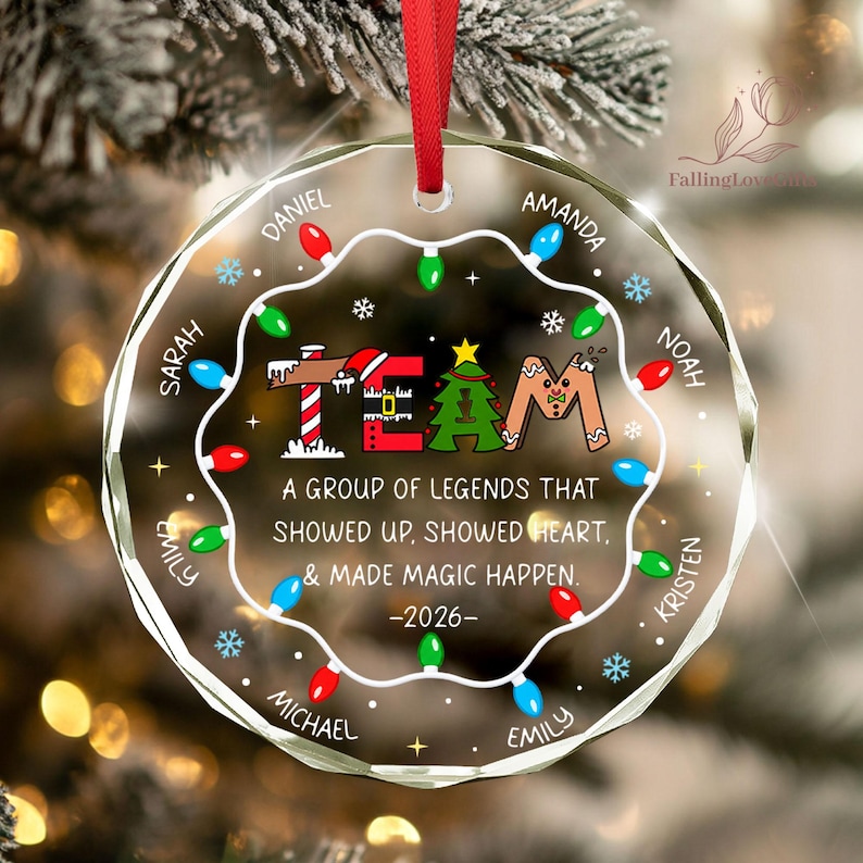 May include: A round, clear glass Christmas ornament with a red ribbon hanger. It features a string of colorful holiday lights, snowflakes, and the text "TEAM" with festive icons. It includes the quote "A GROUP OF LEGENDS THAT SHOWED UP, SHOWED HEART, & MADE MAGIC HAPPEN. -2026-" and several names printed around the edge.