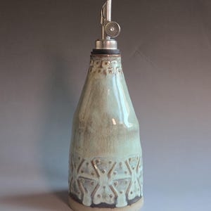 May include: A ceramic olive oil bottle with a stainless steel spout. The bottle has a light green glaze with a geometric pattern near the base. The spout is angled and has a small, round cap. The bottle is approximately 9 inches tall.