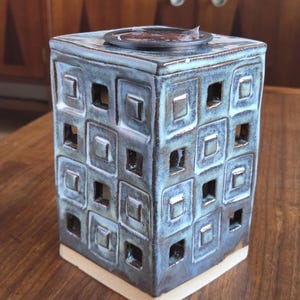 May include: A square teal ceramic candle holder with a geometric square pattern and cut-out designs. The candle holder has a cream-colored base and a dark metal top. The teal glaze has a mottled appearance.