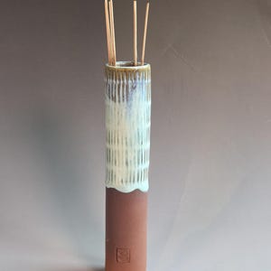 May include: A tall, cylindrical ceramic incense holder. The bottom is a reddish-brown color, transitioning to a white and light blue textured pattern. Several incense sticks are visible inside the holder.