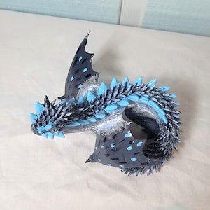 Könnte beinhalten: Eine kleine, handgefertigte Drachenfigur mit einem schwarzen und silbernen Körper, blauen Flügeln und einem blauen und silbernen Schwanz. Der Drache ist in einer defensiven Position zusammengerollt.