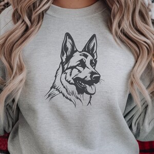 May include: A gray sweatshirt featuring a black and white graphic of a German Shepherd dog's head. The dog is depicted in profile, with its tongue hanging out. The sweatshirt is a neutral color, suitable for various outfits.