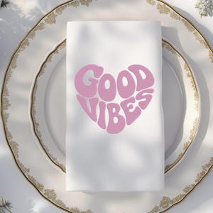 May include: A white cloth napkin with a pink heart-shaped design that reads "GOOD VIBES." The napkin is placed on a white plate with a gold rim. The design is in a retro font.