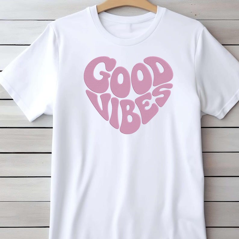 May include: White t-shirt with a pink heart-shaped graphic that reads "GOOD VIBES" in a retro font. The shirt has short sleeves and a crew neck, perfect for casual wear.