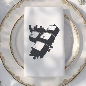 Peut inclure: Serviette blanche pli&eacute;e avec un motif noir repr&eacute;sentant les touches WASD d'un clavier d'ordinateur. La serviette est pos&eacute;e sur une assiette blanche avec un bord dor&eacute;. Design moderne et minimaliste.