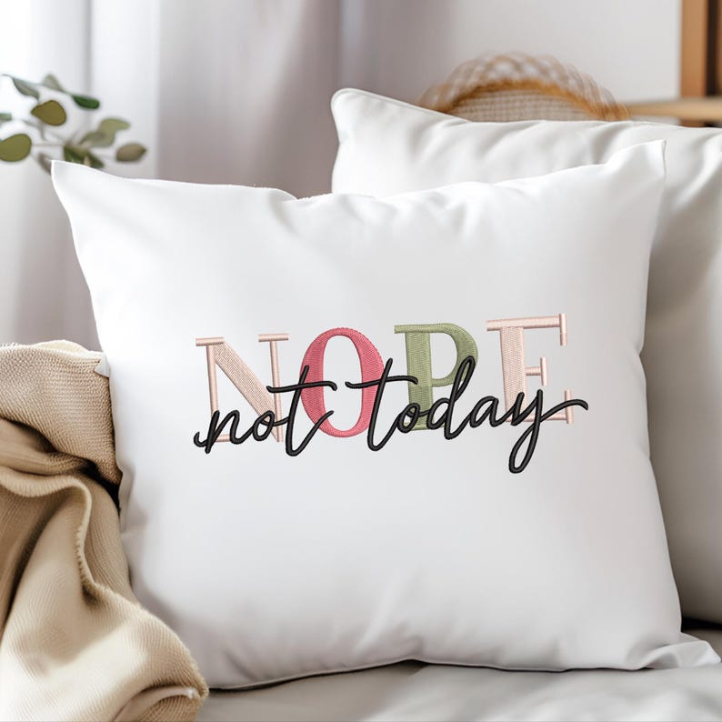 May include: White square pillow with the embroidered text "NOPE not today" in pink, green, and black. The pillow is on a white surface, with a neutral-colored blanket visible.