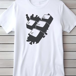 Peut inclure: T-shirt blanc avec un motif noir repr&eacute;sentant les touches de clavier W, A, S et D. Les touches sont regroup&eacute;es avec un fond sombre &eacute;clabouss&eacute;. Le t-shirt est fait d'une mati&egrave;re douce.